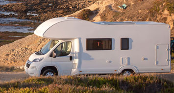 A moving white caravan in the middle of a road beside the shoreline projecting a caravan insurance.